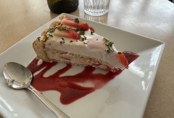 A slice of strawberry cake with topping and syrup on a square white plate with a spoon on the side.
