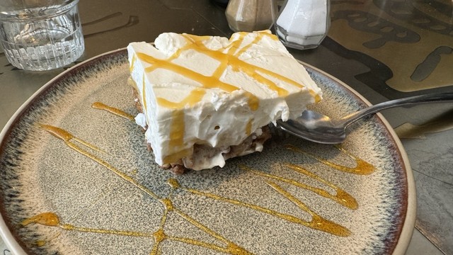 A slice of pie with whipped cream topping drizzled with a yellow sauce on a ceramic plate, next to a glass of water and eating utensils.