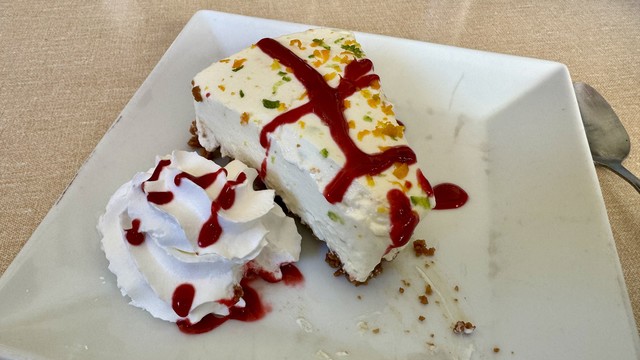 Une tranche de cheesecake avec de la crème fouettée et du coulis de fruits sur une assiette blanche carrée.